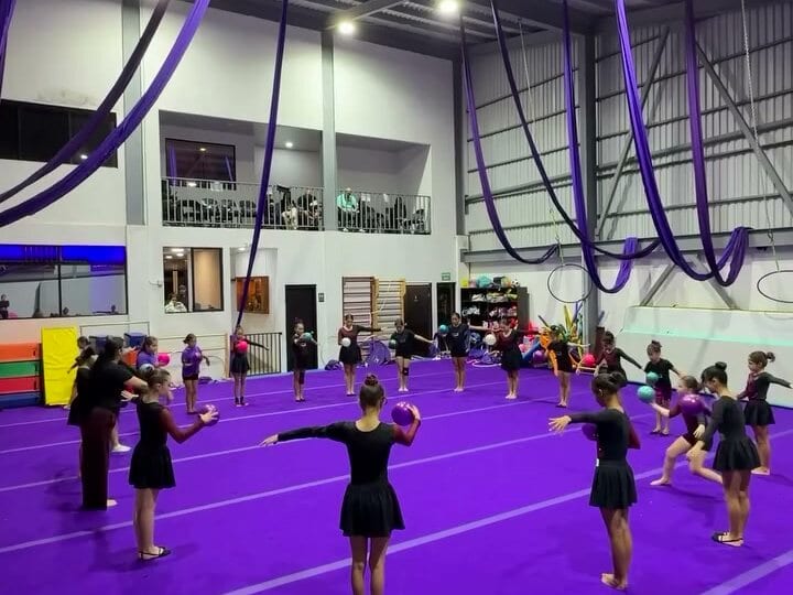 Clase de gimnasia rítmica sobre el piso violeta, con las telas aéreas colgadas del techo